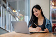 © bongkarn - A woman taking notes in a book while studying online or working remotely on her laptop at a cafe.