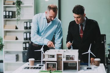 Engineers meeting about wind turbine model and house model. Renewable energy and housing development concept. Team of architects and engineers in modern office discussing design structure.