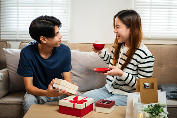 Wall Mural - A young man and woman are sitting together on a couch, exchanging gifts with happy expressions. Surrounded by festive decorations.