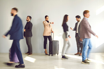 Young woman with a traveling suitcase standing by the wall and looking at her watch while waiting at the train station or airport among a blurred crowd of people walking by fast