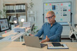 © Tj - Senior businessman gesturing in frustration during a video conference on his laptop, struggling with technical issues or a difficult conversation in a busy office environment