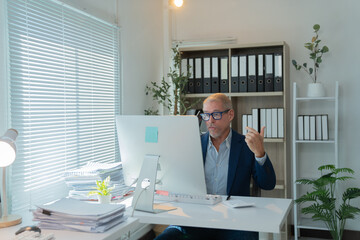 Senior businessman wearing suit and glasses is sitting at his office desk having a video conference on his desktop computer, explaining something with hand gestures