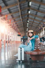 A young woman in casual attire and hat sits on a bench at a train station, with a suitcase beside her, ready for travel.