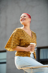 Wall Mural - A cheerful bald woman holds a coffee cup while smiling in a sunny urban environment.