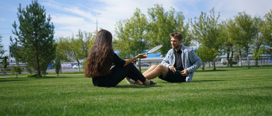 A man and woman sit on the grass in a park, engaged in a lively conversation under a clear blue sky. The atmosphere is relaxed and cheerful, ideal for quality time together.