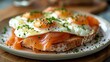© nurionstudio - Close up of a plate of toast topped with smoked salmon, fried eggs, chives, and black pepper.
