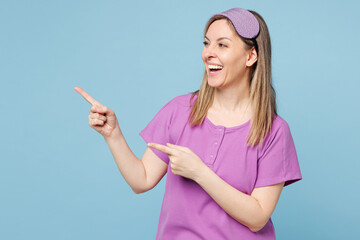 Wall Mural - Calm young woman wears purple pyjamas jam sleep eye mask rest relax at home point index finger aside indicate on area isolated on plain pastel light blue cyan background. Good mood night nap concept.