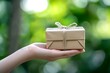 © Marina Shvedak - Hand holding a small zero waste gift box wrapped in brown paper and tied with string on a blurred green bokeh background