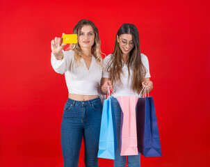 Shopping concept, two beautiful caucasian women standing over red background. Blonde girl showing credit card smiling to camera. Brunette lady holding and looking into paper package bags. Copy space.