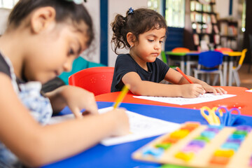 Two Latina girls coloring in a kindergarten classroom, surrounded by scissors, educational games, and coloring supplies