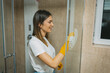© Milos - Wearing yellow gloves, a cheerful woman uses a cloth to clean a shower's glass door, embodying attention to detail and the importance of sanitation.