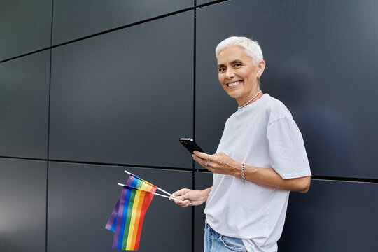 Joyful woman waves pride flag while texting in modern urban space.