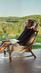 Young male freelancer seated on a wooden chair with laptop on his lap, relaxes working remotely in a peaceful outdoor setting surrounded by nature. Work-life balance