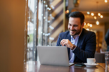 A Caucasian male executive engages with his laptop at a cafe table, dressed in business attire with coffee and smartphone nearby, reflecting a blend of professionalism and casual work environment.