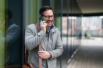 Mid adult Caucasian businessman in smart casual attire engaged in a phone conversation, standing by a modern urban backdrop, reflecting technology integration in corporate lifestyle.