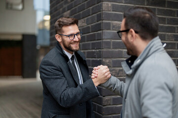 A smiling mid adult Caucasian businessman in a suit shakes hands with another professional man, signifying a meeting or agreement, against an urban backdrop.