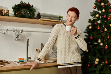 A young man dressed warmly savors coffee beside a beautifully decorated Christmas tree.