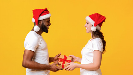 Wall Mural - Christmas tradition. Loving multiracial couple in Santa hats exchanging Xmas gifts, congratulating each other with winter holidays, yellow background