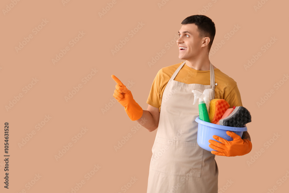 Young man with cleaning supplies pointing at something on beige background