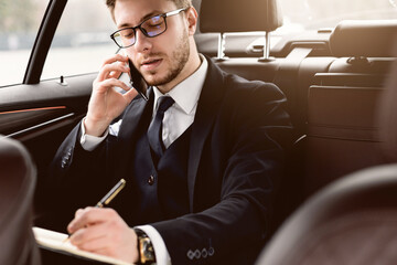 Always In Touch Concept. Young manager in glasses writing in notebook talking on smart phone sitting in back seat of car