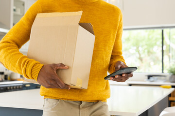 African american man holding cardboard box and using smartphone at home, multitasking efficiently