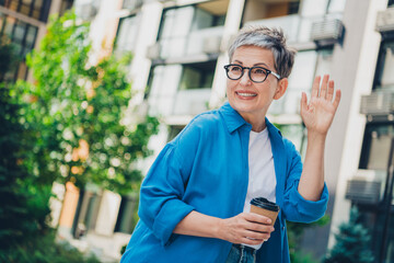 Photo of nice aged lady hold coffee wave hi wear blue shirt downtown street city outdoors