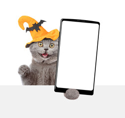 Poster - Surprised kitten wearing hat for halloween holds shows smartphone with white blank screen in it paw above white blank board. Empty space for text. Isolated on white background
