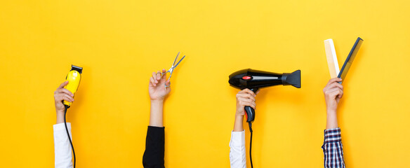 Hands holding hair cutting tools including hair dryer, combs, scissors and clipper in isolated yellow color banner background