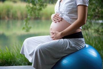 Pregnant woman holding belly sitting on fitness ball in nature