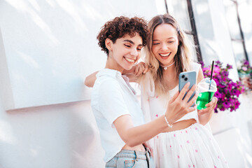 Two young beautiful smiling trendy female. Carefree women posing in street. Positive models having fun. Cheerful and happy. Hold smartphone, look at mobile cellphone screen, use phone apps