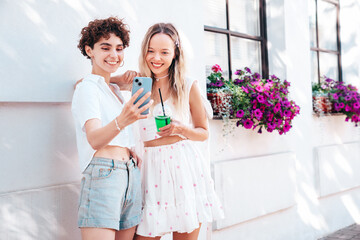 Two young beautiful smiling trendy female. Carefree women posing in street. Positive models having fun. Cheerful and happy. Hold smartphone, look at mobile cellphone screen, use phone apps