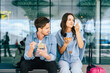 © Iryna - Young adults enjoying snacks while waiting at airport terminal. Two young adult friends, a Caucasian man and woman, eating snacks while sitting on the bench at airport terminal, outdoor.