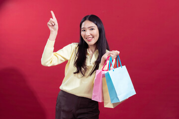 Asian woman holding colorful shopping bags in one hand, pointing upward with the other hand, smiling brightly. Dressed in a yellow top and brown pants against a red background