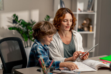 in the children's room a schoolboy in a white sweater and a blue checkered shirt at desk does homework mom explains the task