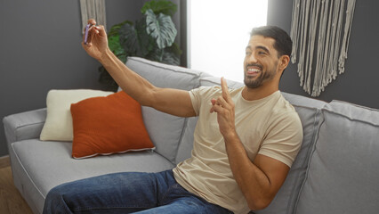 Young hispanic man with beard smiles sitting indoors at home taking selfie in modern living room on sofa making cheerful gesture