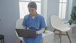 © Krakenimages.com - A focused hispanic woman in blue medical scrubs reviews data on a laptop in a bright, modern clinic room.