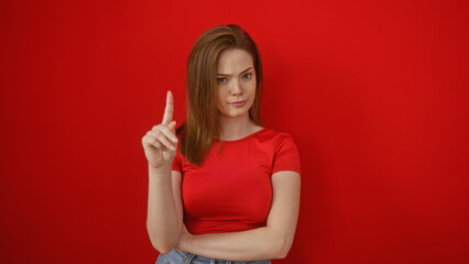 Young woman with red hair and in red shirt against a red wall making a gesture with her finger raised, looking confident, attractive and isolated over the bright background