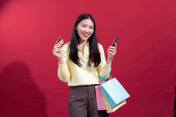 Asian woman smiling, holding a credit card and a smartphone, with colorful shopping bags on her arm. Wearing a yellow top and brown pants, standing against a red background