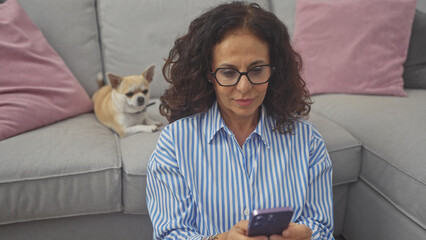 Sticker - A middle-aged hispanic woman uses a smartphone in a cozy living room with a chihuahua dog resting on the couch beside her.