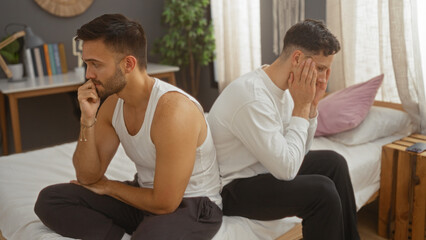 Two men sitting on a bed in a bedroom looking distressed and upset, representing a gay couple experiencing relationship issues at home, with a cozy and intimate interior setting