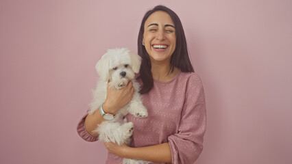 A joyful young hispanic woman in a pink sweater hugs a fluffy white bichon maltese dog against a pink wall.