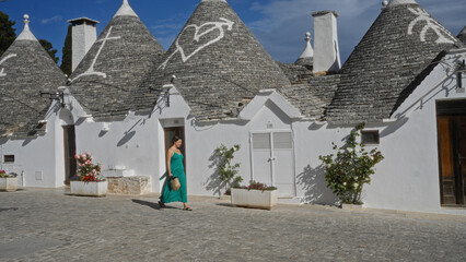 Wall Mural - A young hispanic woman in a vibrant dress strolls down a charming street in alberobello, italy, lined with iconic trulli houses under a clear blue sky.