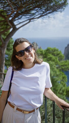 Poster - Woman smiling outdoors in capri, italy, wearing sunglasses and white shirt, standing near railing with sea and trees in the background, radiating happiness and relaxation.