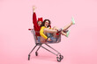 © kegfire - Two joyful friends having fun and laughing while sitting in a shopping cart against a pink background.