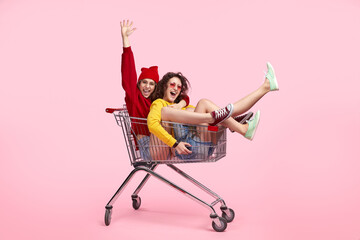 Two joyful friends having fun and laughing while sitting in a shopping cart against a pink background.