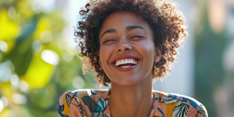 A woman with curly hair is smiling and wearing a floral dress. She has her eyes closed and is looking at the camera