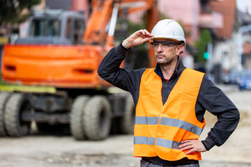 Construction worker in safety gear analyzing site conditions while machinery operates in the background during daylight hours