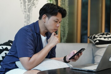 Asian man sitting on living room sofa with pillow in lap holding smartphone reading social network and thinking fun playing game