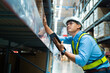 © KANGWANS - A man in a safety vest is looking at a tablet while standing in a warehouse. He is wearing a hard hat and he is focused on the tablet