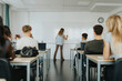 © Maskot - Rear view of teacher writing on whiteboard while male and female students sitting in classroom